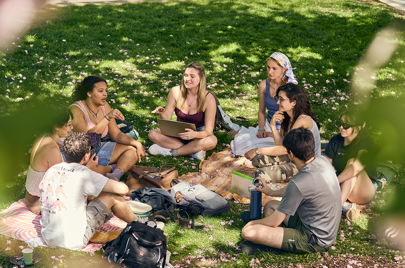 Eight students with books and backpacks sitting in a circle on the Quad and talking to each other