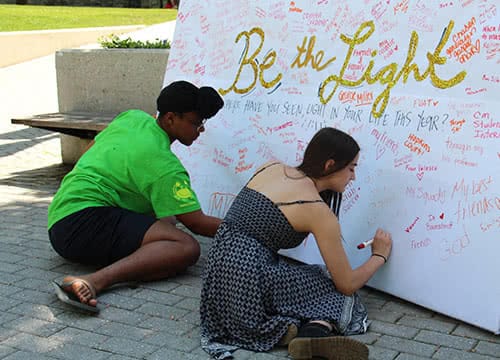 Students sitting on the ground writing a message on a board that says Be the Light"