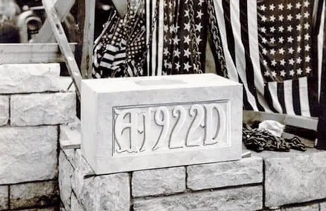 A black and white photo of a cornerstone with A.1922.D carved into it, with American flags in the background