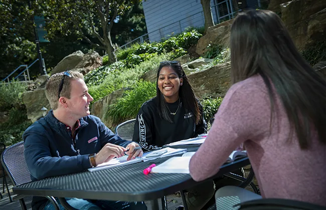 Three students talk and smile outside at a table
