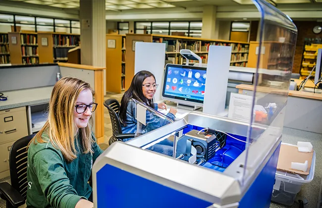 Two students working with a 3D printer in the library