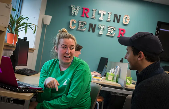 Two students talking. The text "Writing Center" can be seen on the wall in the background