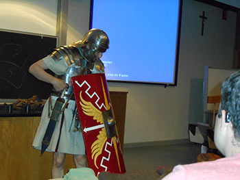 Matt Amy in armor, with a sword and shield, in a classroom