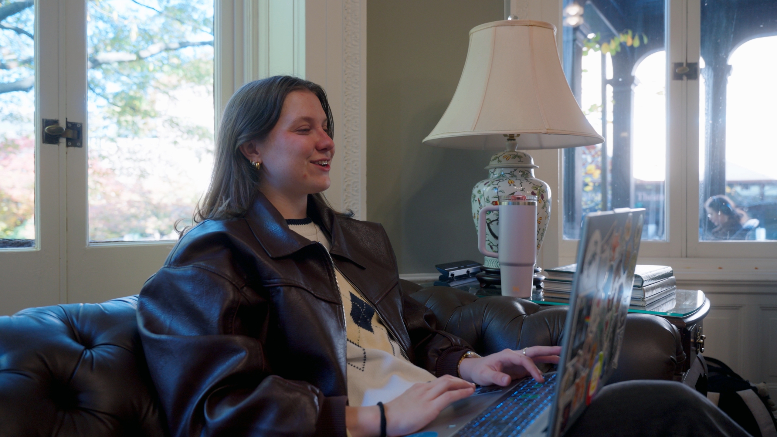 A person sits on a couch using a laptop, smiling, with a lamp, books, and window in the background.