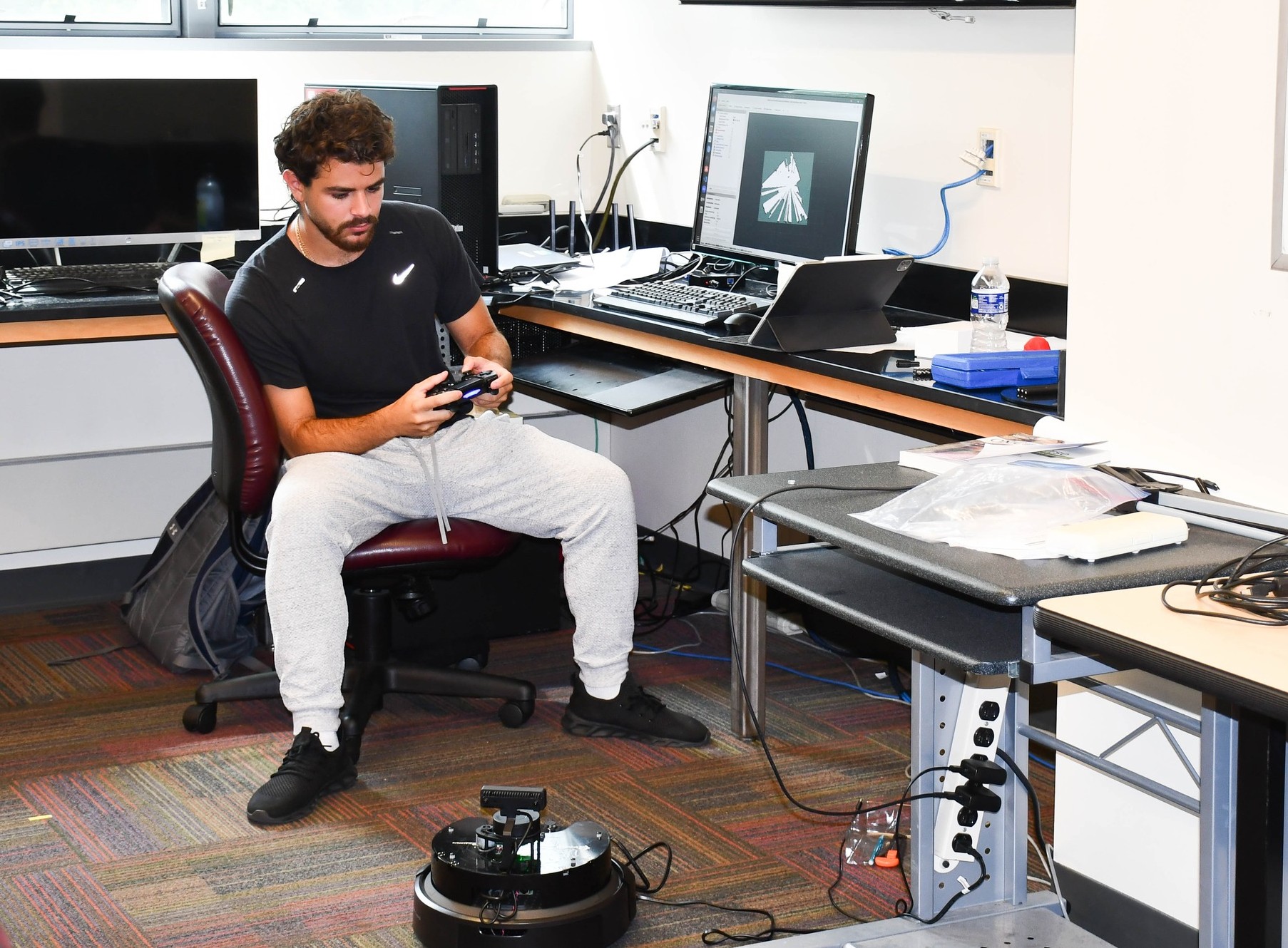 A student sits at a desk and controls a round robotic device on the floor using a controller.