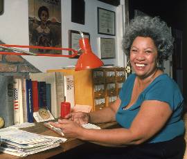 Toni Morrison wears a blue shirt and sits at a desk with a red lamp and books