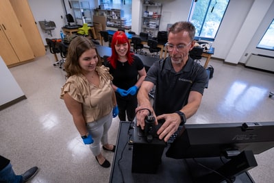 Two students wearing latex gloves observe while professor positions forensics equipment