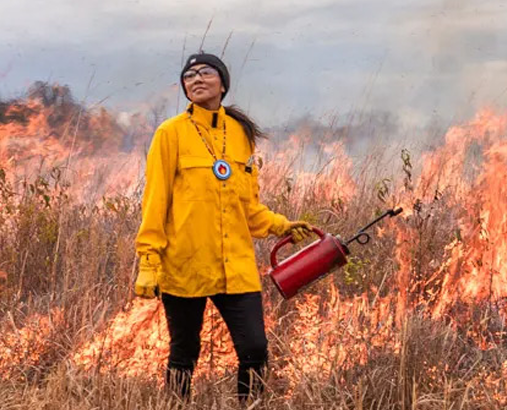 A photograph of a woman standing in a field on fire.
