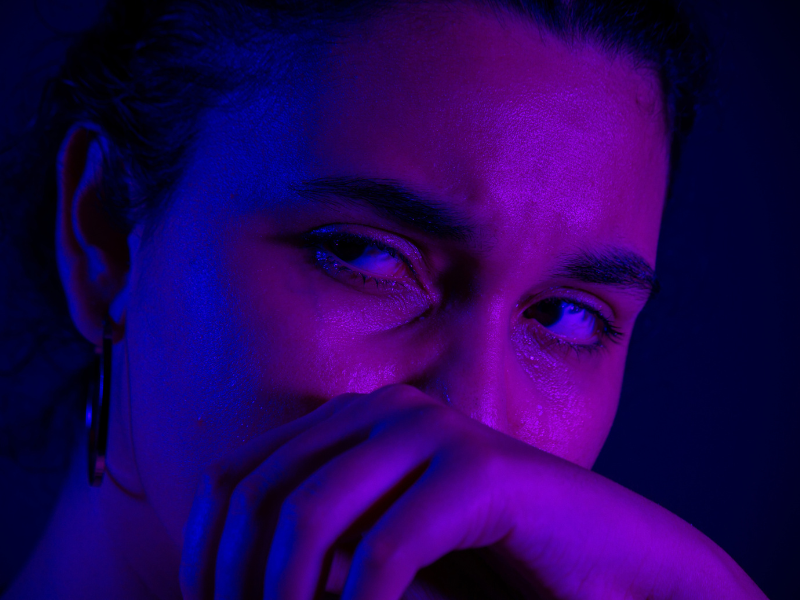 Close-up portrait of a person covering their mouth with one hand under blue and magenta lighting.