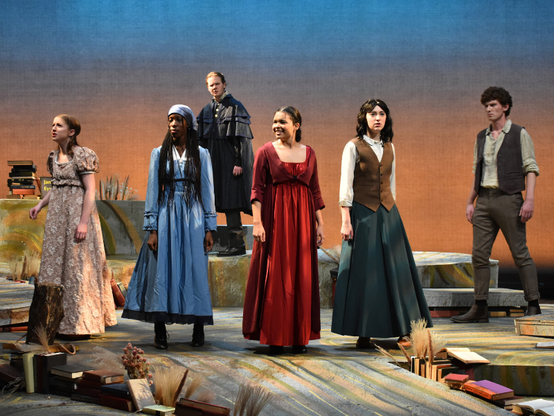 A theatrical stage scene shows six actors in period-style clothing standing across a tiered, book-filled set beneath a blue-to-brown backdrop.