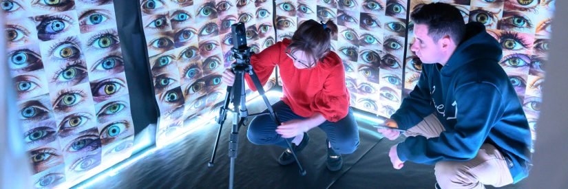 Two people working with a camera in a room filled with images of eyes on the walls.