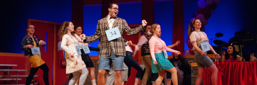 People on stage performing a choreographed dance, wearing signs with numbers.
