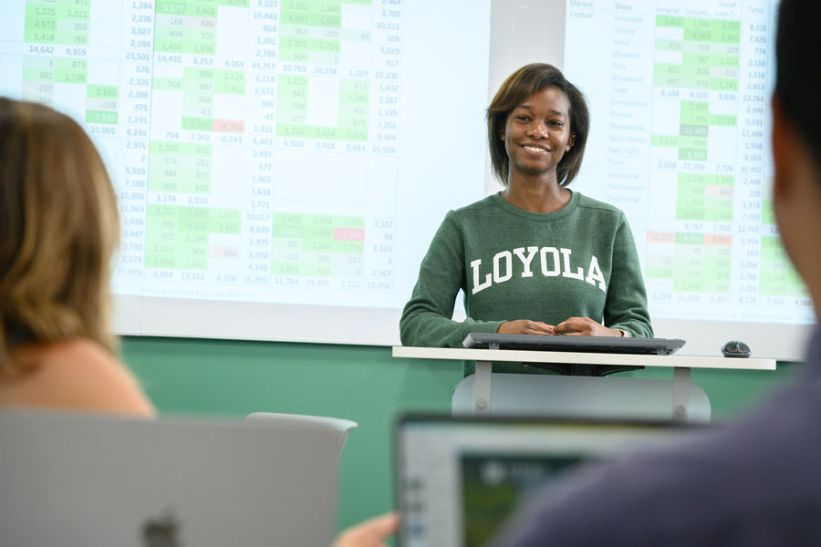 In a modern classroom, a presenter with spreadsheets on the screen behind her.