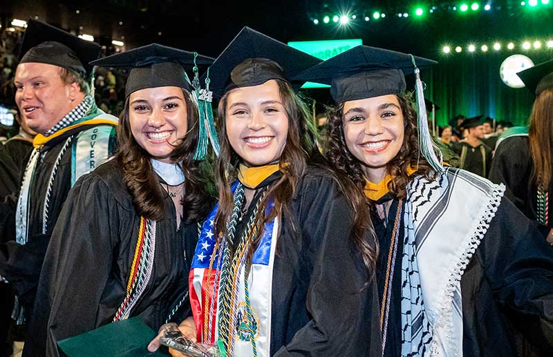 Three students with big smiles in graduation caps and gowns and heavily decorated with honors