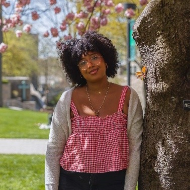 Naomi smiles and leans against a cherry blossom tree on the grassy quad