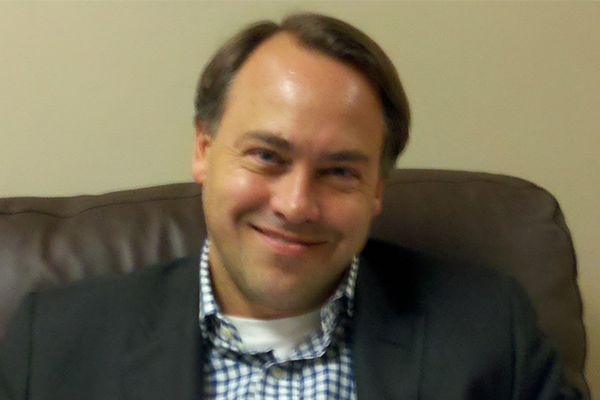 Man in checked shirt and dark sport coat sitting in desk chair