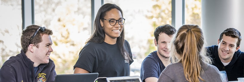 5 students smiling while doing group work in a bright, naturally lit room in the library 