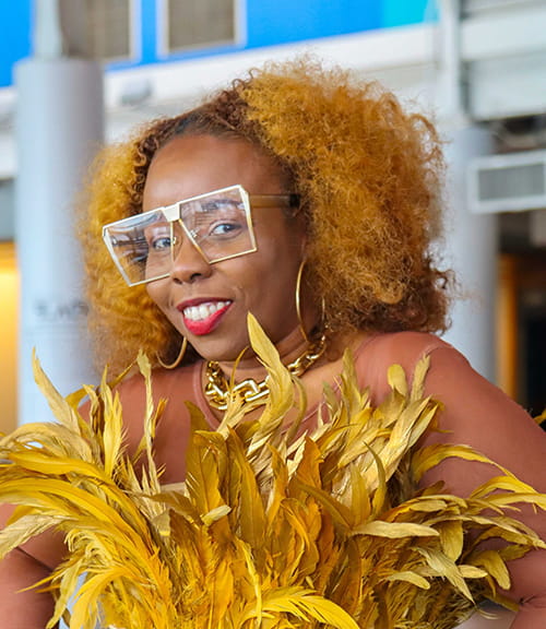 Ms. Teekay smiles wearing a gold feather top with stylish square glasses.