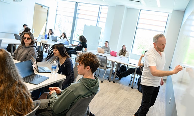 A professor writes on a whiteboard in a classroom with clustered seating arrangements