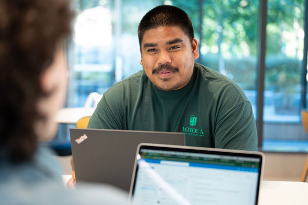 A graduate student in a green Loyola shirt works at a table with a fellow student in the Fernandez Center.