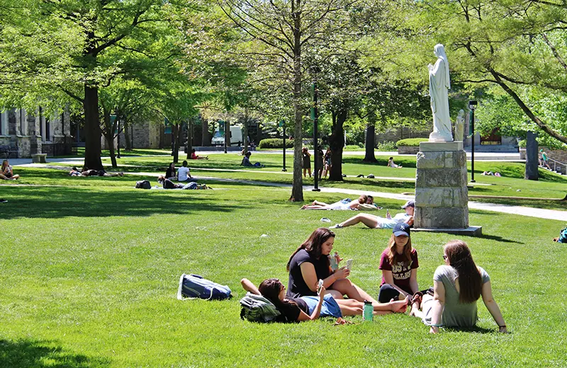 A statue of Mary watches over the grassy green quad on a sunny day as students are scattered all around