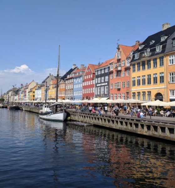 Nyhavn, the city center in copenhagen, an ocean inlet with colorful buildings lining the water with lots of people eating outside.