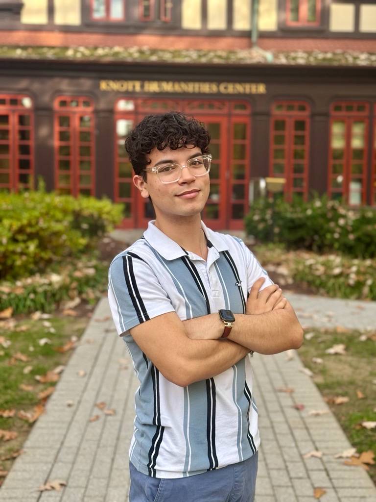 Juan standing with his arms crossed across his chest, smiling at the camera as he stands in front of the Humanities building.