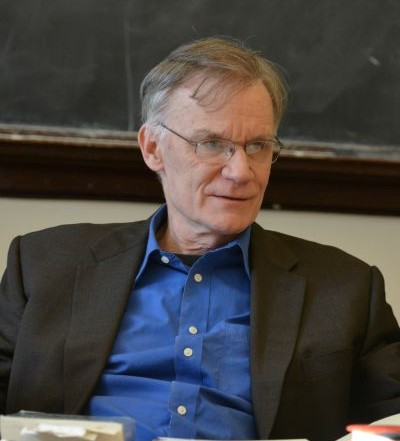 man in blue shirt and dark jacket sitting in front of chalkboard