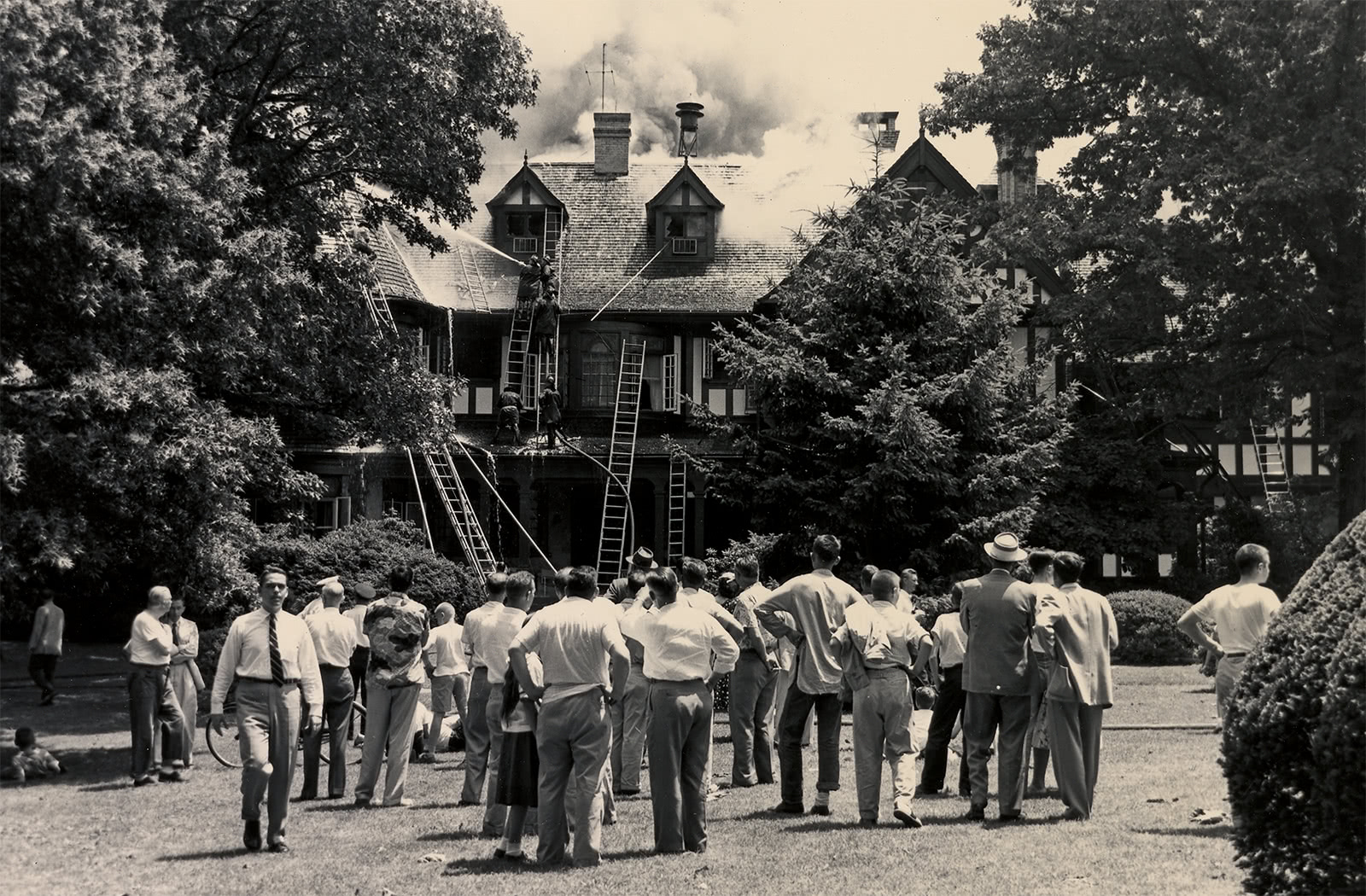 Black and white photo of a large group of people watching firefighters extinguish a fire in the Humanities Building