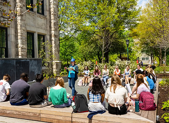 A group of students sitting on benches outdoors on a bright and sunny day watching a lecture