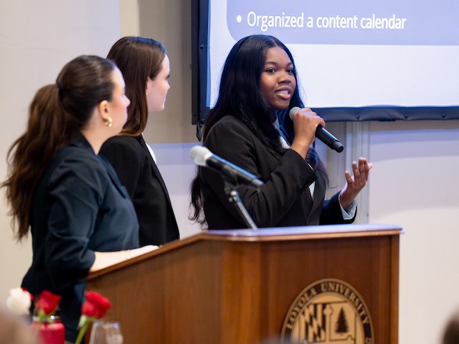 Three women at a lectern with a projection screen behind them. One is speaking into a microphone.