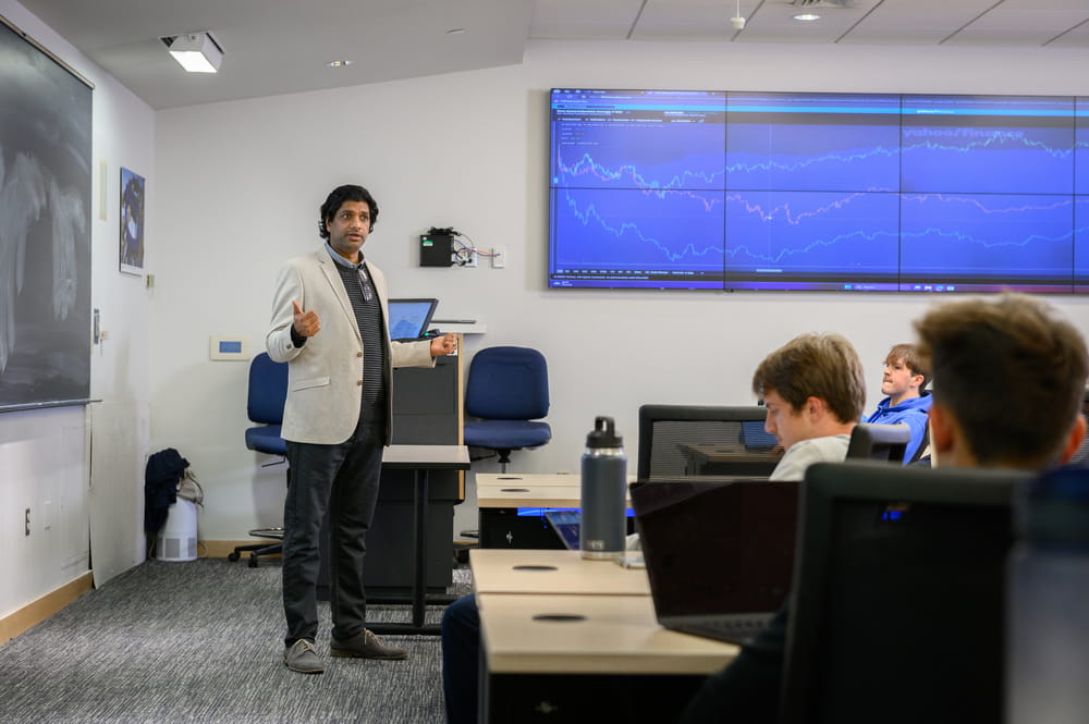 A professor stands at the front of a classroom, teaching a room full of students at their desks