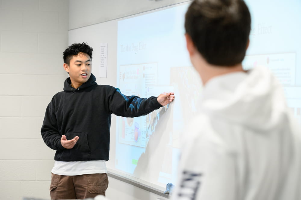 A student stands at the front of a classroom and presents a project to a classmate while pointing to information on a projected screen