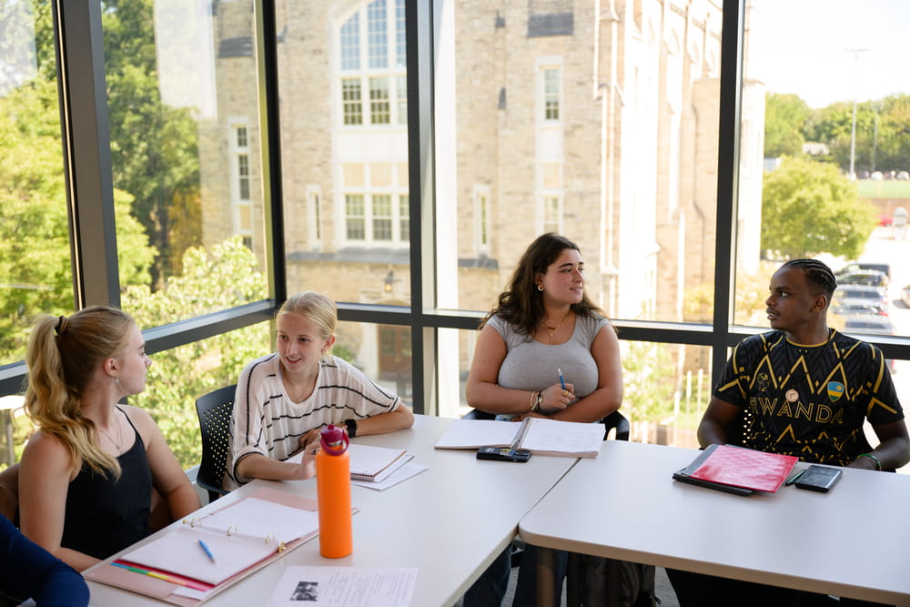 Four students sit together around a table during  class in the Fernandez Center, talking and working with their class materials