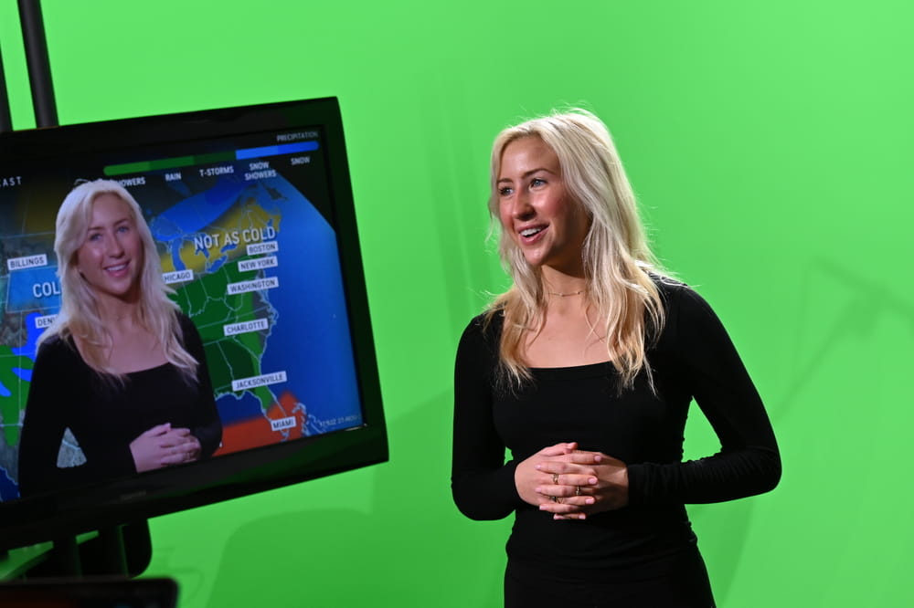 A student on a film set, between a video monitor and a green screen.