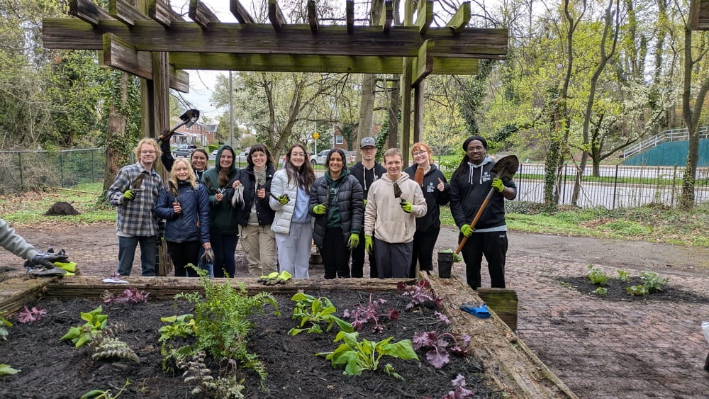 A group of students pose with gardening equipment behind a raised flower bed in Evesham Park.