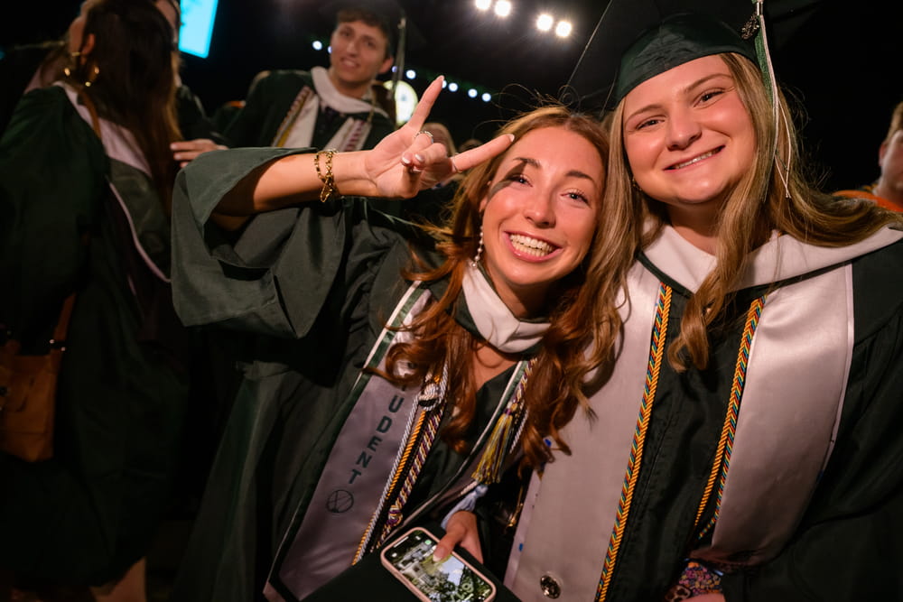 Two students in graduation regalia smile at the camera