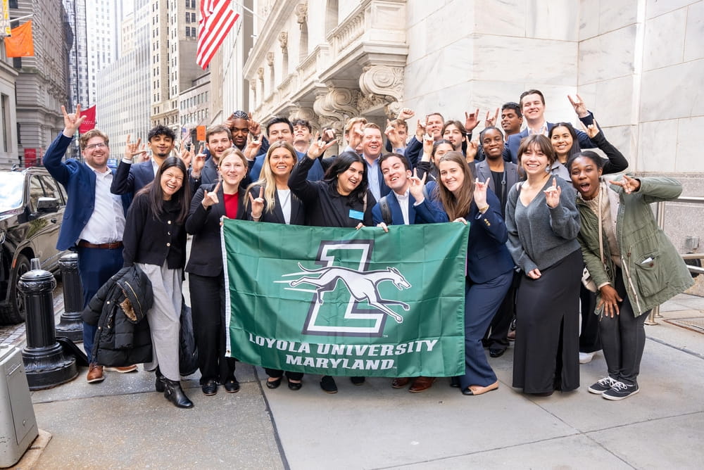 A large group of students poses with a Loyola University Maryland flag in front of the New York Stock Exchange