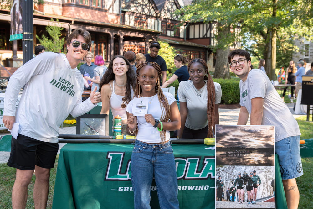A group of students smile for the camera at a table full of promotional materials for the Loyola rowing team