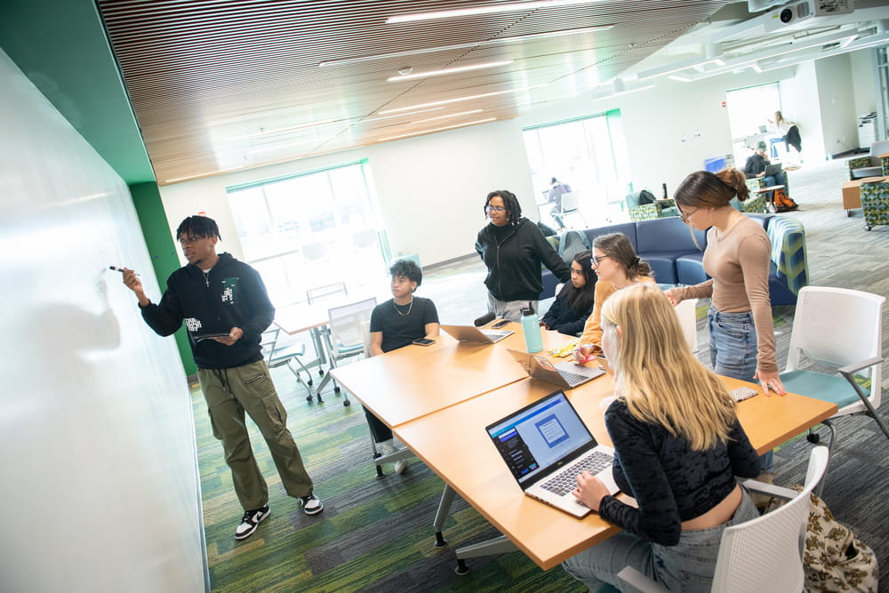 A group of students gathers around a table in the Thrive Center while one student writes on a whiteboard and others  work on laptops