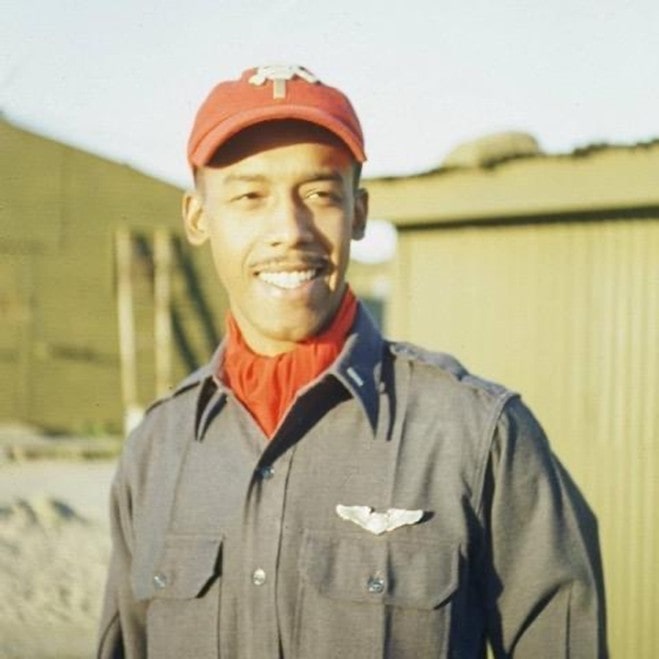 A smiling Black man in gray and red Air Force uniform stands outside with miliary buildings behind him