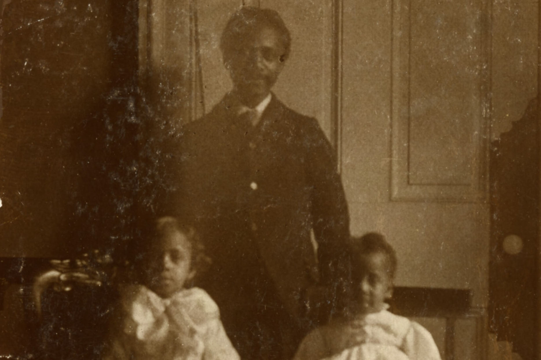 A sepia portrait of a Black man dressed in a suit standing behind two Black children dressed in white and seated on chairs.