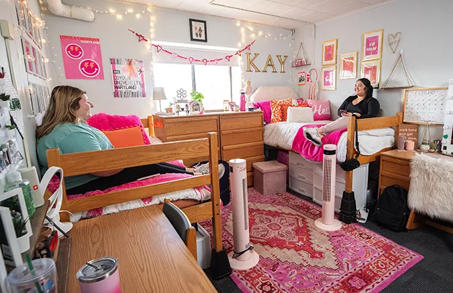 Two students sit on their beds in a room with a bright pink and red rug
