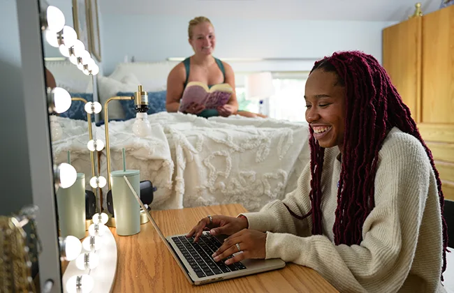A student working on a laptop, while her roommate reads a book in the background