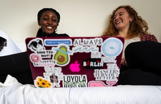 Two students sit on a bed looking at a laptop and laughing