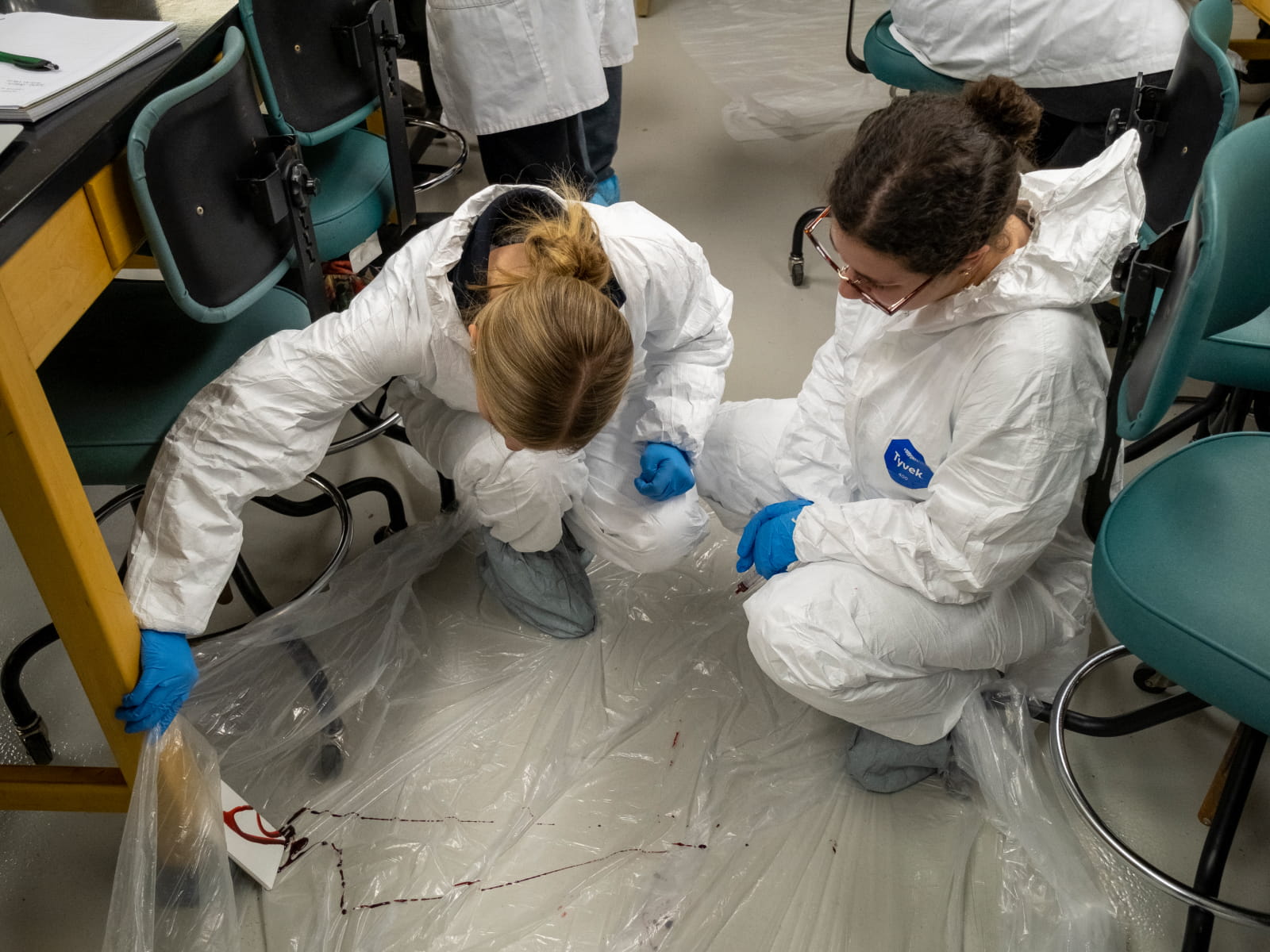Two students in white protective coveralls and gloves inspect the floor