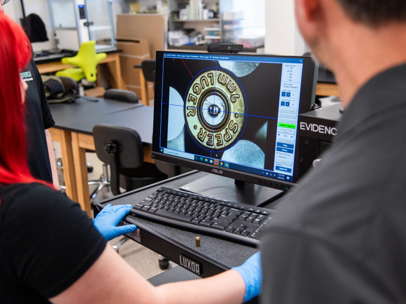 A forensic science graduate student wearing gloves looks at a bullet under a microscope on a computer screen.