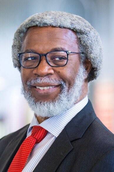 A guy with a grey hair, smiling, wearing eyeglasses, red tie, and black coat