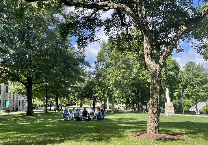 A circle of students meets for class under leafy trees on the Academic Quadrangle. 