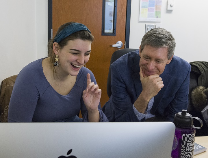 A student works on a laptop while a professor smiles and looks on.
